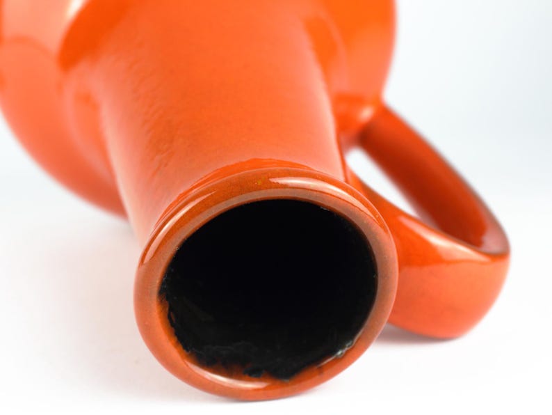 May include: Close-up of a vibrant orange ceramic vase with a glossy finish. The vase has a wide opening and a handle. The interior is black. The design is simple and modern, suitable for home decor.