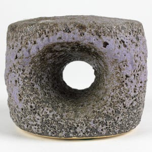 Può includere: Una scultura in ceramica grigio scuro con un foro al centro. La scultura ha una superficie ruvida e testurizzata e una smaltatura viola chiaro.