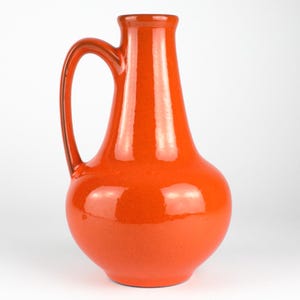 May include: A vibrant orange ceramic pitcher with a curved handle. The pitcher has a bulbous base and a slender neck, with a glossy finish. The pitcher is approximately 10 inches tall.