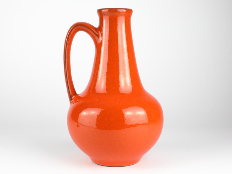 May include: A vibrant orange ceramic pitcher with a curved handle. The pitcher has a bulbous base and a slender neck, with a glossy finish. The pitcher is approximately 10 inches tall.