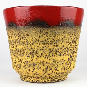 Può includere: Un vaso di ceramica con una vivace combinazione di colori. Il bordo superiore è rosso lucido, che sfuma in un corpo giallo maculato con macchie marroni scure. Il vaso ha una forma leggermente svasata e una superficie strutturata.