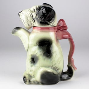 Op de afbeelding: Een zwarte en witte keramische theepot in de vorm van een hond met een roze strik op zijn kop. De theepot heeft een tuit en een handvat in de vorm van een hondenstaart.