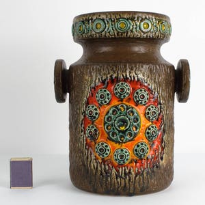 以下が含まれることがあります： テクスチャ加工された表面とカラフルな幾何学模様が施された茶色のセラミック花瓶。花瓶は口が広く、側面に小さな取っ手が2つ付いています。