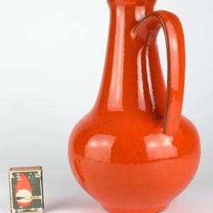 May include: A glossy, orange ceramic pitcher with a curved handle. The pitcher has a rounded base and a narrow neck. A matchbox with the word "SOLO" is next to the pitcher.