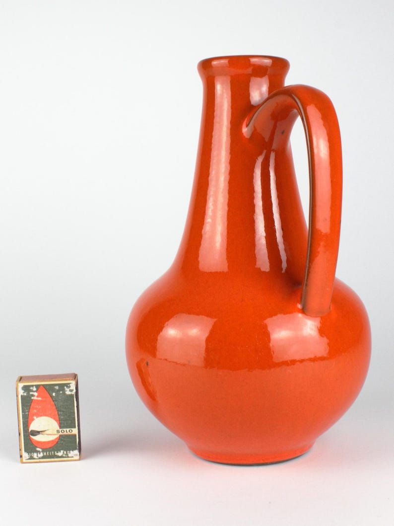 May include: A glossy, orange ceramic pitcher with a curved handle. The pitcher has a rounded base and a narrow neck. A matchbox with the word "SOLO" is next to the pitcher.