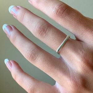 May include: A silver ring with a simple, rectangular design. The ring is worn on the ring finger of a hand.