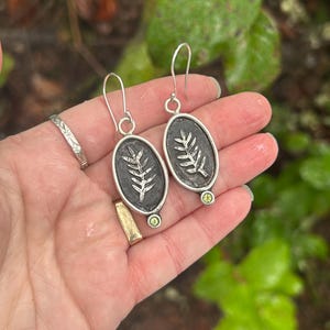 May include: Silver oval earrings with a dark background and a raised leaf design. Each earring has a small, round, light green stone at the bottom. The earrings are held up by a silver hook.