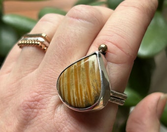 Op de afbeelding: Zilveren ring met een grote, druppelvormige steen met bruine en beige strepen. De ring heeft een klein gouden accent. Een andere gouden ring is zichtbaar op de achtergrond.