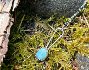 May include: A silver pendant necklace with a light blue, triangular stone. The pendant is set in a silver frame and hangs from a delicate silver chain. The necklace is displayed on a bed of green moss and small rocks.