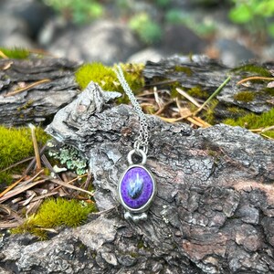 May include: A silver necklace with a pendant featuring a purple stone with blue and black inclusions. The pendant is oval-shaped and set in a silver bezel. The necklace is displayed on a textured, natural wood surface.