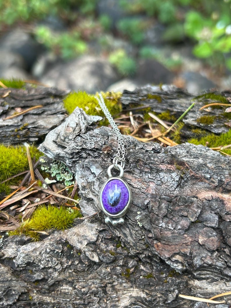 May include: A silver necklace with a pendant featuring a purple stone with blue and black inclusions. The pendant is oval-shaped and set in a silver bezel. The necklace is displayed on a textured, natural wood surface.