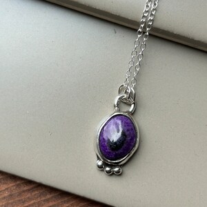 May include: A silver necklace featuring an oval pendant with a vibrant purple stone and a dark center. The pendant is framed in silver and has three small silver beads at the bottom. The chain is delicate and silver.