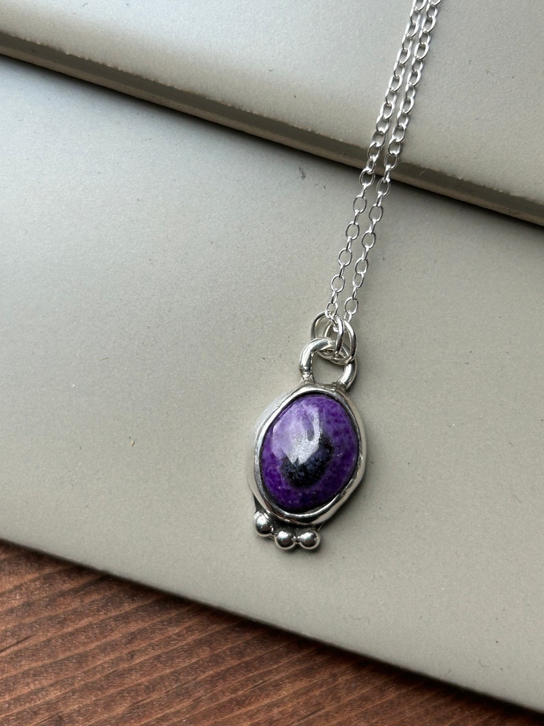 May include: A silver necklace featuring an oval pendant with a vibrant purple stone and a dark center. The pendant is framed in silver and has three small silver beads at the bottom. The chain is delicate and silver.