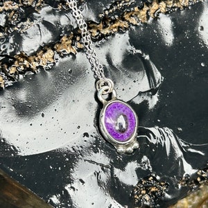 May include: A silver necklace with a pendant featuring a purple stone. The oval-shaped stone is set in a silver bezel and hangs from a delicate silver chain. The pendant has a small, decorative silver design at the bottom.