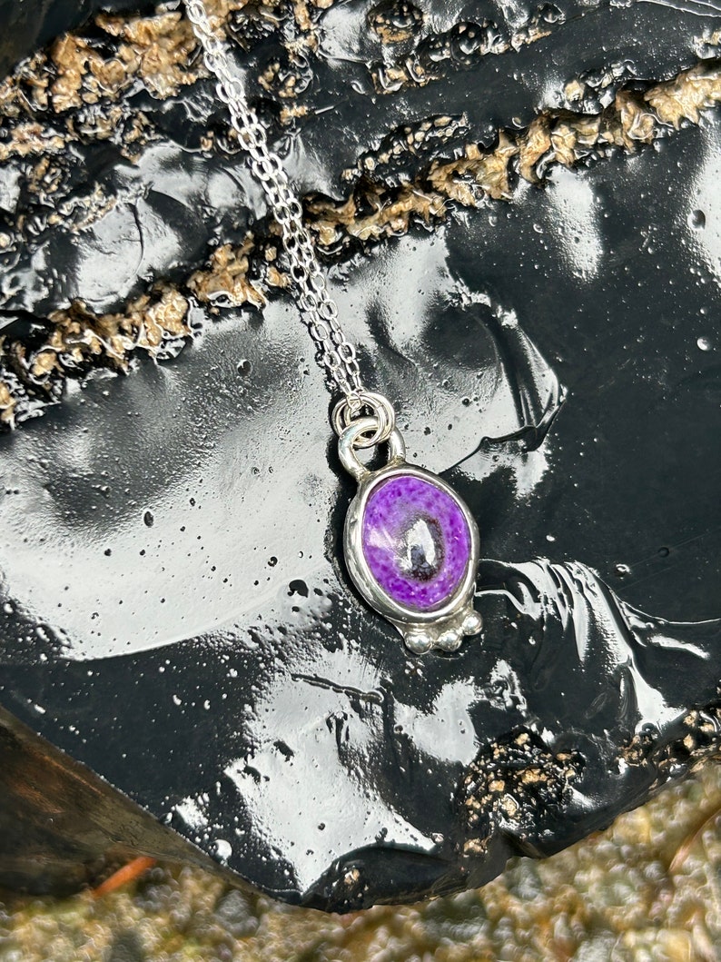 May include: A silver necklace with a pendant featuring a purple stone. The oval-shaped stone is set in a silver bezel and hangs from a delicate silver chain. The pendant has a small, decorative silver design at the bottom.