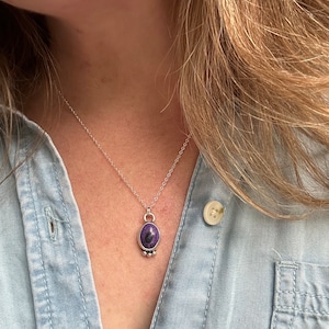 May include: Silver necklace with a pendant featuring a purple stone with dark inclusions. The oval-shaped stone is set in a silver bezel and hangs from a delicate silver chain. The necklace is worn over a light blue denim shirt.