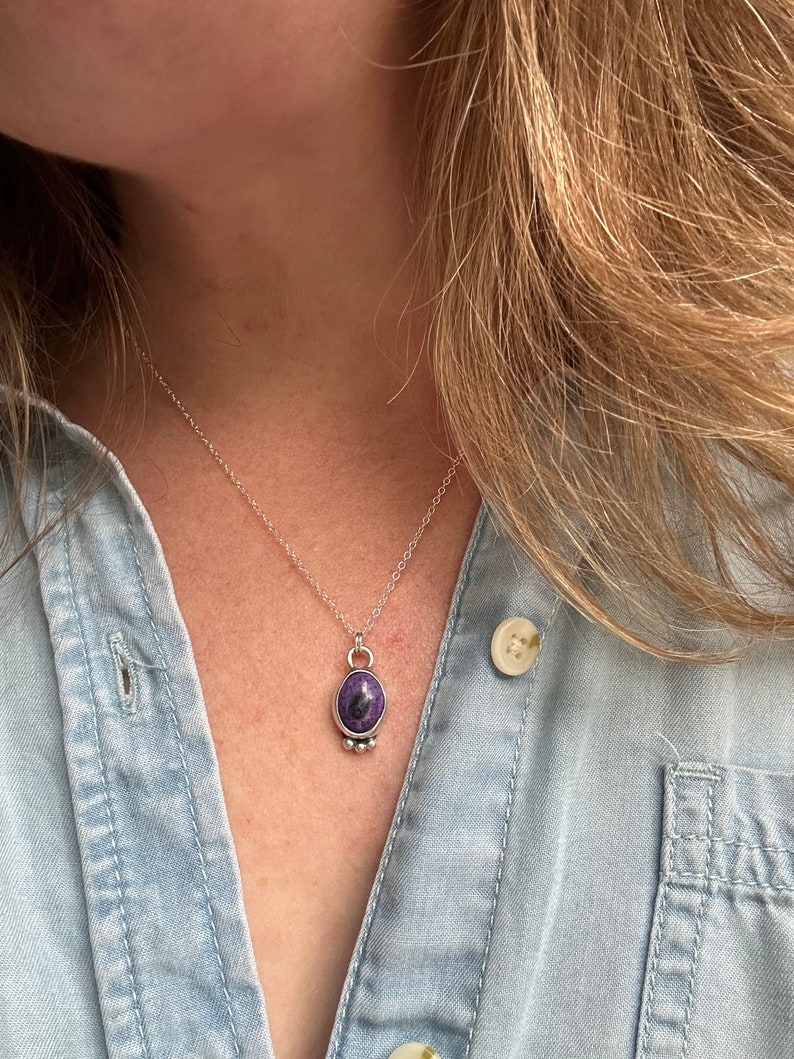 May include: Silver necklace with a pendant featuring a purple stone with dark inclusions. The oval-shaped stone is set in a silver bezel and hangs from a delicate silver chain. The necklace is worn over a light blue denim shirt.