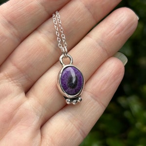 May include: A silver pendant necklace featuring a purple stone with black inclusions. The oval-shaped stone is set in a silver bezel with three small silver beads at the bottom. The necklace has a delicate silver chain.