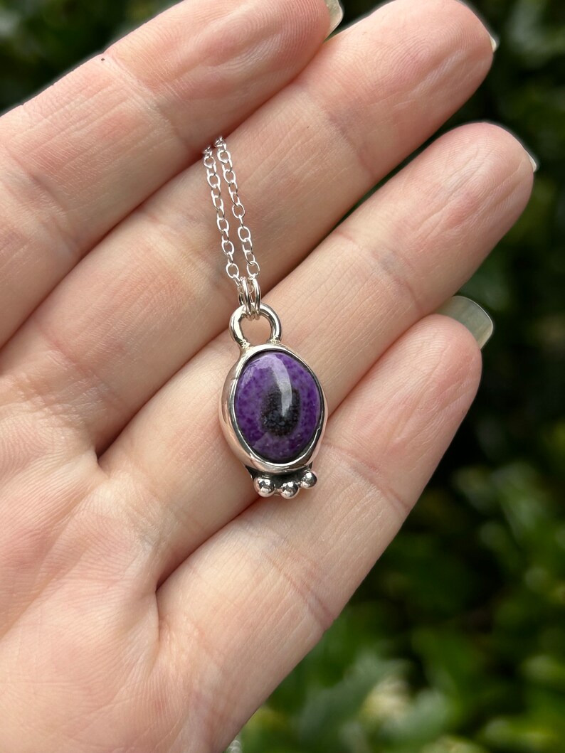 May include: A silver pendant necklace featuring a purple stone with black inclusions. The oval-shaped stone is set in a silver bezel with three small silver beads at the bottom. The necklace has a delicate silver chain.