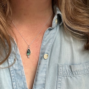 May include: A silver necklace with a teardrop-shaped pendant. The pendant features a dark green stone with black inclusions and a small silver bead at the bottom. The necklace is worn over a light blue denim shirt.