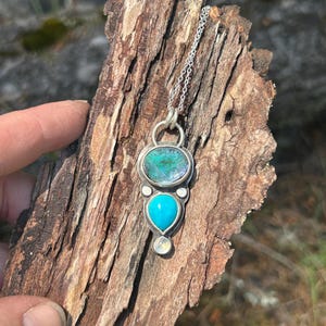 May include: A silver pendant necklace featuring an oval-shaped iridescent stone above a turquoise teardrop-shaped stone. A small, round, white stone is below the turquoise stone. The pendant hangs from a delicate silver chain.