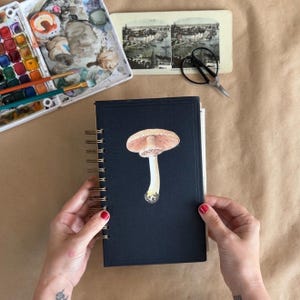 May include: A black spiral-bound notebook featuring a centred illustration of a light brown mushroom with a detailed cap and stem. The notebook is positioned near a watercolour paint set, a vintage black and white photograph, and black scissors.