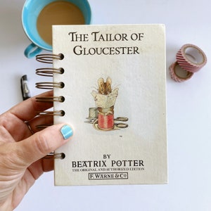 Może przedstawiać: Spiralny notatnik z tytułem "The Tailor of Gloucester" autorstwa Beatrix Potter. Okładka przedstawia ilustrację myszy czytającej gazetę. Niebieski kubek kawy i ozdobna taśma znajdują się w tle.
