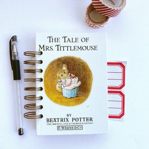 Peut inclure: Un carnet à spirales avec la couverture de "The Tale of Mrs. Tittlemouse" de Beatrix Potter. Un stylo transparent et deux rouleaux de ruban adhésif décoratif sont également visibles. Le livre présente une illustration de souris.
