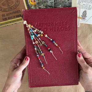 May include: A deep red hardback book titled Arthur Mee's Hero Heroes featuring a decorative bookmark attachment. The bookmark consists of multiple strands of colourful glass and metal beads, including small charms shaped like keys and bees.