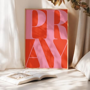 May include: A framed art print with the word "PRAY" in large, bold letters. The letters are pink against a red background. The print is in a wooden frame, and a book is open in front of it.