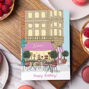 Puede incluir: Una tarjeta de felicitación rectangular con una colorida ilustración de la fachada de una pastelería con un toldo rosa que dice Naomi's Cake Shop & Cafe, mesas de bistró al aire libre y el texto Happy Birthday impreso en rosa en la parte inferior.