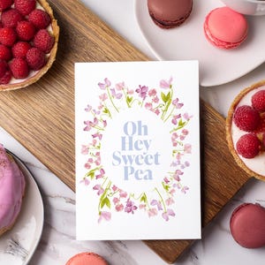 May include: A rectangular white greeting card featuring a watercolor illustration of pink and purple sweet pea flowers arranged in an oval frame. The center of the card displays the blue text "Oh Hey Sweet Pea" in a clean, sans-serif font.