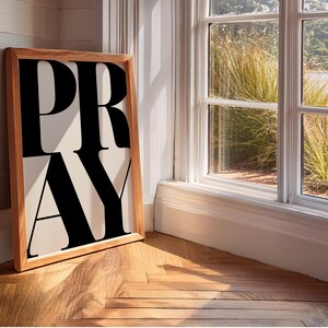 A framed print featuring the word "PRAY" in large, black, bold letters on a cream background. The frame is light-coloured wood. The statement christian artwork leans against a white wall next to a window overlooking a garden.
