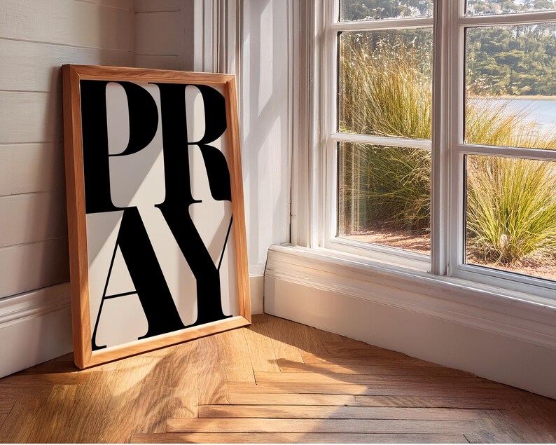 A framed print featuring the word "PRAY" in large, black, bold letters on a cream background. The frame is light-coloured wood. The statement christian artwork leans against a white wall next to a window overlooking a garden.