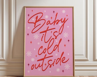 May include: A rectangular wall art print in a light wooden frame featuring a pink background with the red cursive text Baby it's cold outside. The design is decorated with white snowflakes and small red star shapes scattered across the surface.