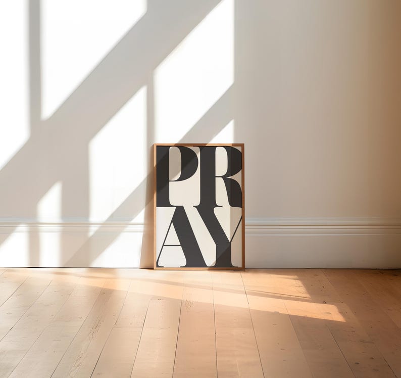 A bold framed print with the word "PRAY" in bold, black letters on a white background. Modern christian typography print for trendy apartments. The frame is a light brown wood and sits in a contemporary loft style gallery space.