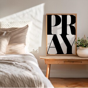 Christian wall art print positioned next to a bed in a cosy, neutral bedroom. Statement wall art poster with bold typography which reads PRAY in oversized all caps letters. Faith wall art printable in modern black and white typography,  for Christian home decor. Digital download for prayer space or bedroom. Printable artwork for religious lifestyle decor.