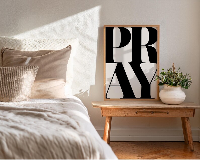 Christian wall art print positioned next to a bed in a cosy, neutral bedroom. Statement wall art poster with bold typography which reads PRAY in oversized all caps letters. Faith wall art printable in modern black and white typography,  for Christian home decor. Digital download for prayer space or bedroom. Printable artwork for religious lifestyle decor.