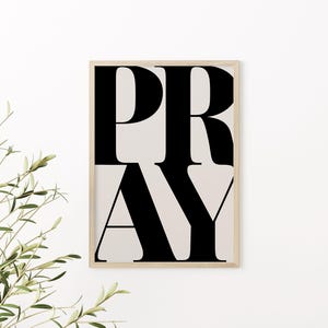 May include: A framed art print with the word "PRAY" in large, bold, black letters. The letters are stacked vertically against a light beige background. The frame is a light wood color, and the print is displayed against a white wall.