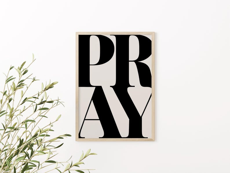 May include: A framed art print with the word "PRAY" in large, bold, black letters. The letters are stacked vertically against a light beige background. The frame is a light wood color, and the print is displayed against a white wall.
