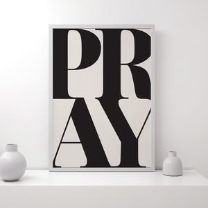 A framed print featuring the word "PRAY" in large, black, bold letters on a cream background. The artwork is displayed on a white shelf with three white ceramic vases. The style is minimalist and modern.