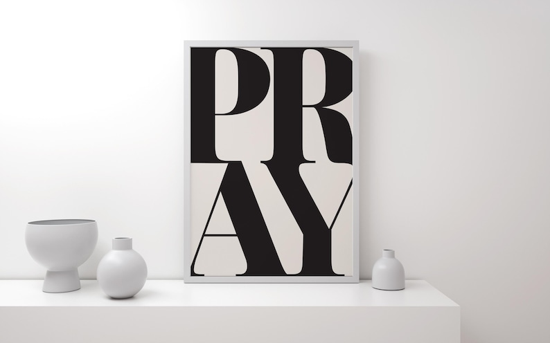 A framed print featuring the word "PRAY" in large, black, bold letters on a cream background. The artwork is displayed on a white shelf with three white ceramic vases. The style is minimalist and modern.