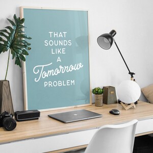 Könnte beinhalten: Ein rechteckiger Wandkunstdruck in einem hellen Holzrahmen mit einem staubblauen Hintergrund und weißem Text, der THAT SOUNDS LIKE A Tomorrow PROBLEM lautet. Der Druck steht auf einer Schreibtischoberfläche neben einem Laptop, einer Lampe und Dekor.