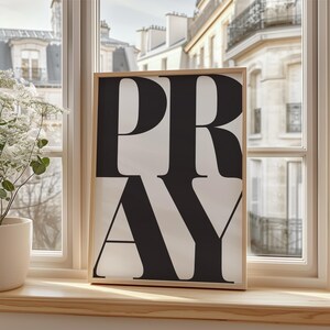 A framed print featuring the word "PRAY" in large, black letters on a white backdrop. The artwork is set on a wooden windowsill, with a view of a European building visible through the window.