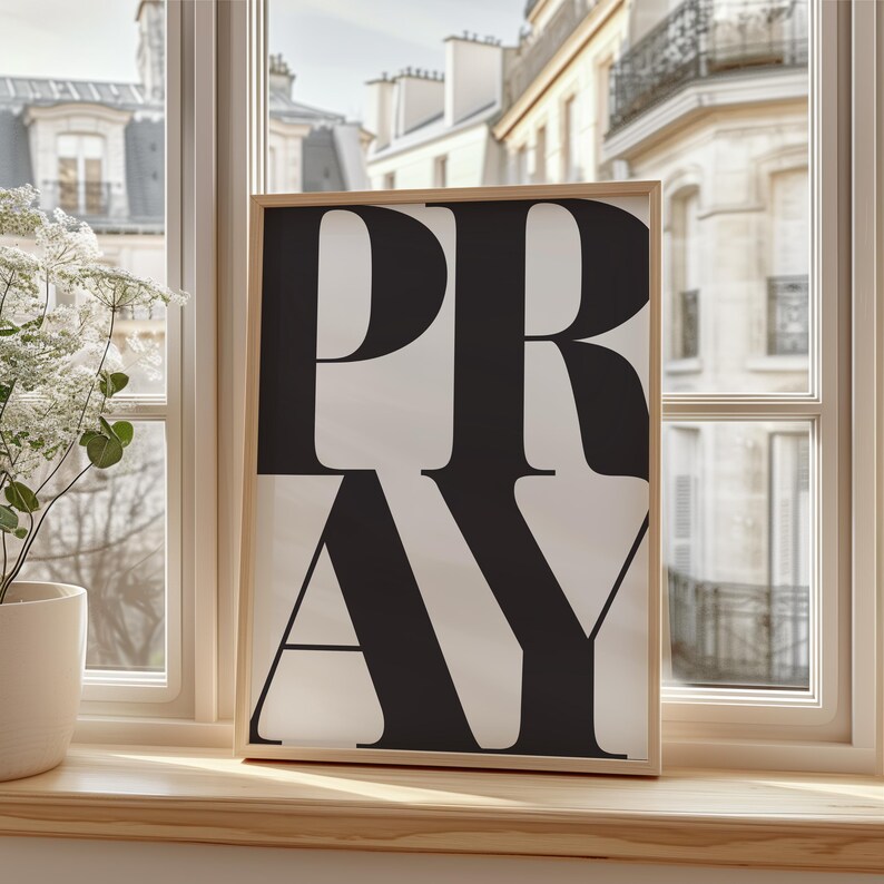 A framed print featuring the word "PRAY" in large, black letters on a white backdrop. The artwork is set on a wooden windowsill, with a view of a European building visible through the window.