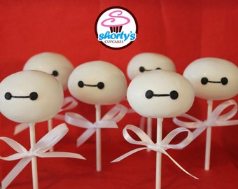 May include: Six white cake pops decorated with black eyes and a black line for a mouth, resembling the character Baymax from the Disney movie Big Hero 6. Each cake pop is on a white stick and has a white ribbon tied around the stick.