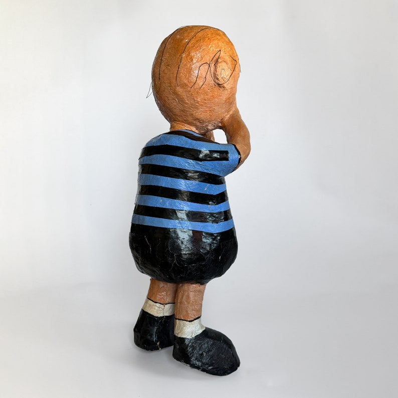 Puede incluir: Una figurita de papel mach&eacute; hecha a mano de un ni&ntilde;o con cabello casta&ntilde;o que lleva una camisa a rayas azules y negras y zapatos negros. La figurita est&aacute; de pie de espaldas a la c&aacute;mara y con la cabeza girada hacia un lado.