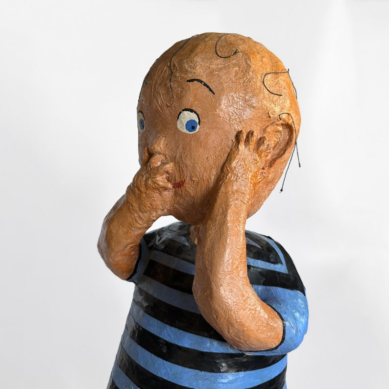 Puede incluir: Una escultura de papel mach&eacute; de un ni&ntilde;o con ojos azules, que lleva una camisa a rayas azules y negras, con las manos cubri&eacute;ndose las orejas y la boca.
