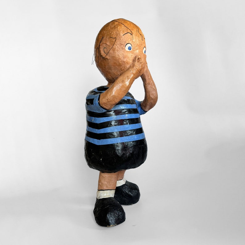 Puede incluir: Una figurita de papel mach&eacute; hecha a mano de un ni&ntilde;o con cabello casta&ntilde;o y ojos azules. Lleva una camisa a rayas azul y negra y pantalones negros. La figurita est&aacute; de pie con las manos cubri&eacute;ndose la boca.