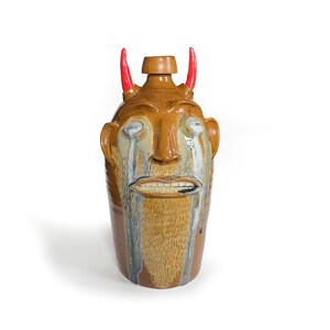 May include: A ceramic jug with a brown glaze and a face sculpted on the front. The face has red horns, a beard, and a sad expression with tears running down its cheeks.